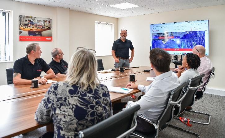 Many people seated round a large table discuss the contents of a screen