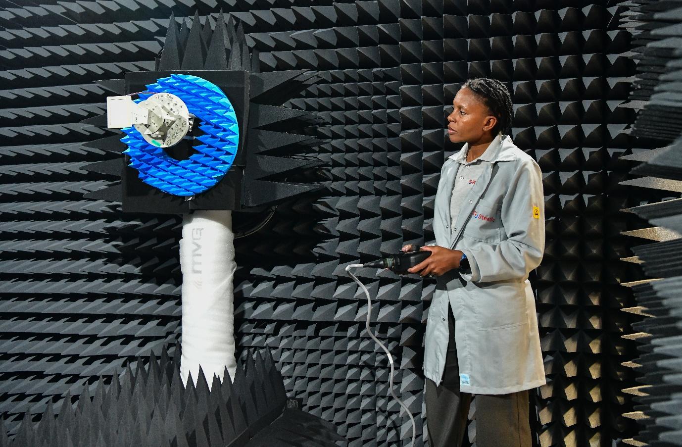 A woman in a lab coat holding testing equipment inside an anechoic chamber, facing a blue antenna mounted on the foam-covered wall.