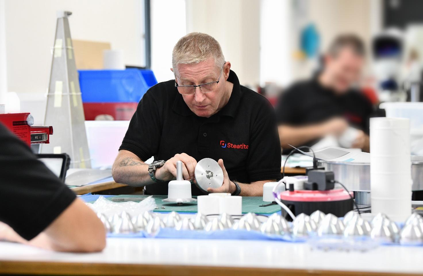 Two men seated at a workbench assemble parts