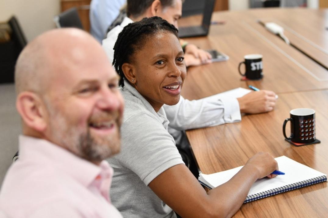 A woman smiling at the camera during a meeting, seated at a wooden conference table with a notebook and pen in front of her; a man in the foreground laughs, and colleagues with a laptop and coffee mugs are visible in the background.