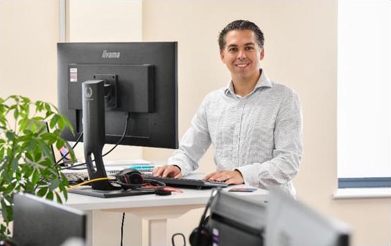 A man smiling at a standing desk with a large monitor in an office.