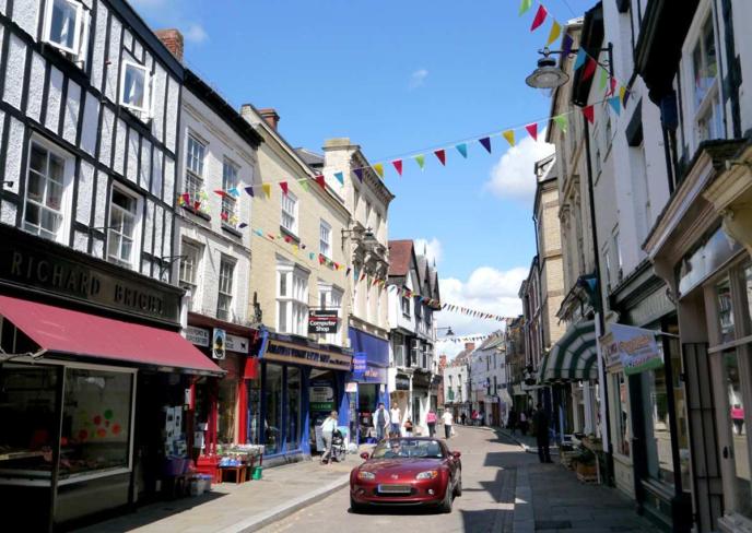 A market town street lined with timber-framed buildings and shops, decorated with colourful bunting, a red convertible car parked in the road.
