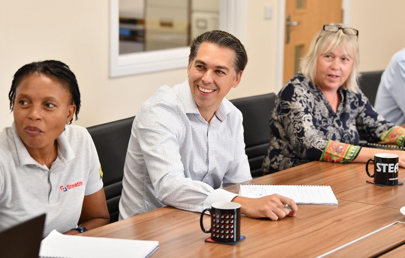 Smiling people at a long table watch a presentation